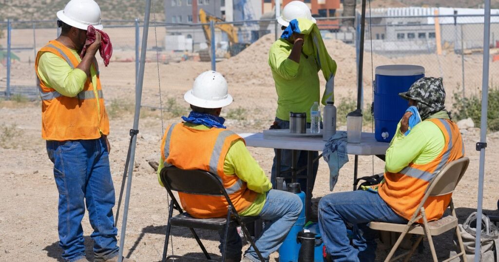 Construction workers in safety hi-vis vests take a hydration and shade break under a portable canopy at a sunny construction site with a water cooler and rest area visible.