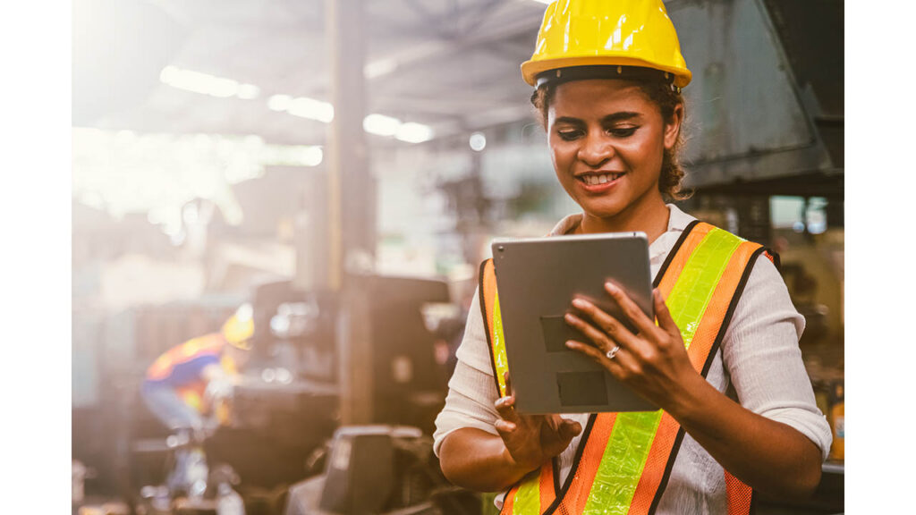 Safety worker in a hard hat and hi-vis vest reviewing a safety observation on a tablet on a factory floor