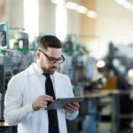 Quality professional in a lab coat reviewing data on a tablet on an industrial workshop floor