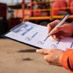 A safety officer in a hard hat writing on a checklist during a risk assessment at an industrial site